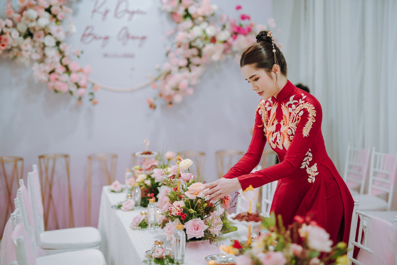 An Asian woman in traditional red dress arranges a floral centerpiece at an elegant indoor event.