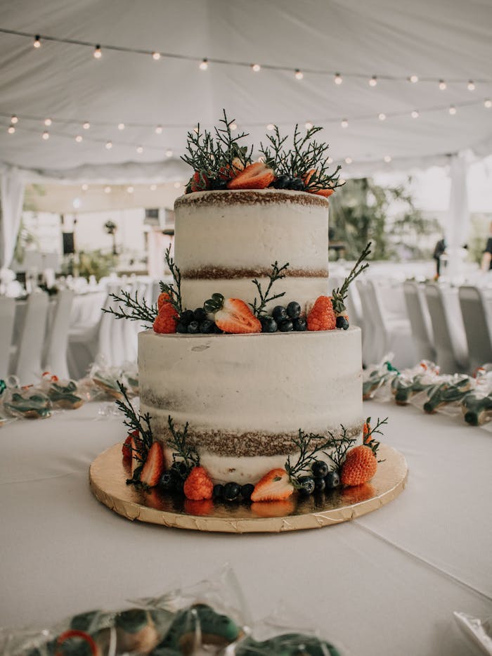Home A two-tier naked wedding cake adorned with fresh strawberries and blueberries in a decorated reception tent.
